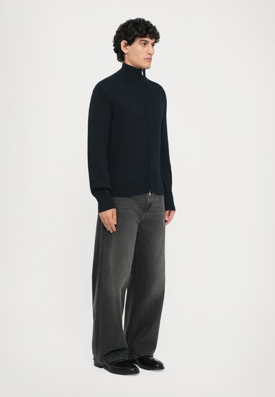 Mörk marinblå ribbad zip-up-tröja med hög krage, parat med löst sittande mörkgrå jeans och svarta skor. Modellen står mot en vit bakgrund.
