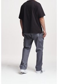 Mann trägt schwarzes T-Shirt, graue Cargo-Hose und weiße Turnschuhe, steht mit dem Rücken zur Kamera vor einfarbigem Hintergrund.