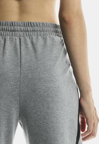 Vedere din spate a unei persoane îmbrăcate cu pantaloni de trening elastici gri deschis, cu o dungi negre, arătând spatele inferior și brațul drept relaxat pe lângă corp.