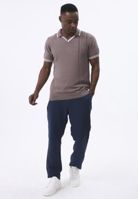 Camisa polo de manga corta marrón con acentos blancos y canalé vertical, combinada con pantalones sueltos azul marino y zapatillas blancas.