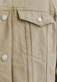 Veste en denim beige avec une poche poitrine, des boutons-pression métalliques et des coutures détaillées sur les cols et les coutures. Tissu doux et texturé.
