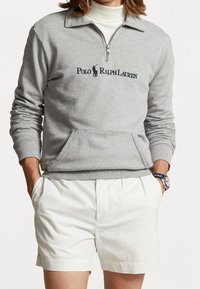 Šedý bavlněný pulover s polo zipem, který má vyšitý logo a přední kapsu. Spárováno s bílými šortkami.