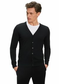 Cardigan nero realizzato in tessuto lavorato a maglia, con scollo a V, maniche lunghe e chiusura con sette bottoni. Texture liscia, abbinato a una camicia bianca.