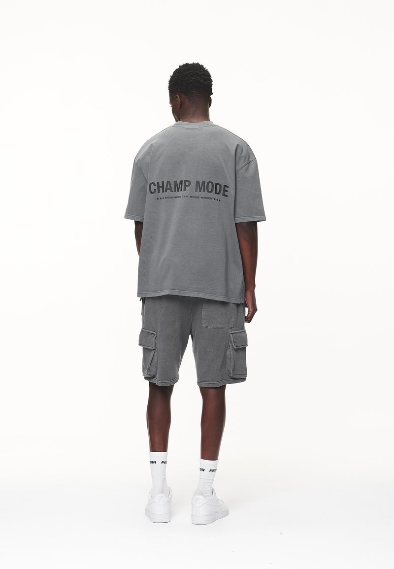 Hombre de pie con la espalda hacia la cámara, vistiendo una camiseta oversized gris con la inscripción "CHAMP MODE", pantalones cortos cargo a juego, calcetines cortos blancos y zapatillas blancas.