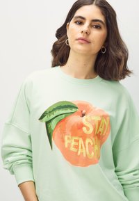 Mätovo zelená mikina s grafickým motívom broskyne a textom "STAY PEACHY" v žltej farbe. Mäkká tkanina s voľným strihom a rukávmi so zúženým lemom.