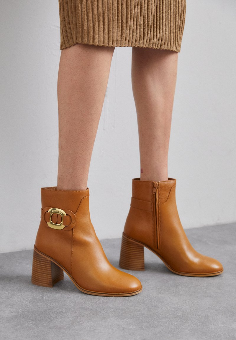 Bottes chelsea en cuir marron avec un talon épais, fermeture éclair sur le côté et une garniture en boucle dorée. Texture lisse avec un design élégant.