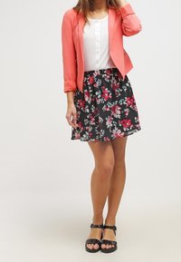 Blazer corallo, top bianco a costine, gonna nera floreale con accenti rosa e sandali neri. Il blazer ha un design aderente e maniche arrotolate.