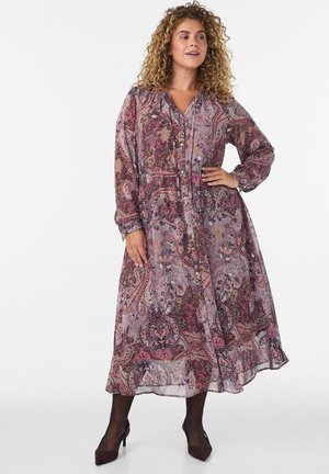 Kvinde med krøllet hår iført en langærmet, ankellang kjole med paisley-mønster og sorte hæle, står med den ene hånd på hoften.