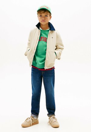 Jongen draagt een beige jas over een groene sweatshirt, blauwe spijkerbroek, beige sneakers en een groene pet met rode letters.