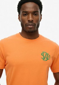 Camiseta de algodón naranja con un logo verde bordado en el lado izquierdo del pecho, cuello redondo y mangas cortas. Textura suave.