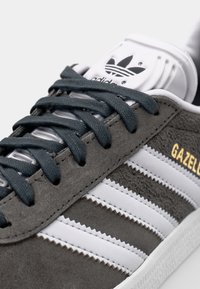 Harmaa mokkanahkakengät, joissa on valkoiset nahkakaistaleet. Varusteena tummanharmaat nauhat ja valkoinen kieli, jossa on kohokuvioitu logo ja kultaiset "GAZELLE"-tekstit.