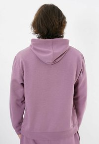 Purple hooded sweatshirt with a soft texture, ribbed cuffs, and hem. Features a relaxed fit and a kangaroo pocket. Rear view shown.