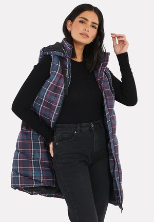 Gilet imbottito con motivo a quadri nei colori navy, bordeaux e grigio. Presenta un cappuccio, zip frontale e tasche laterali, indossato sopra un top nero a coste.