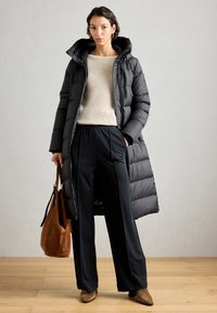Lang zwarte pufferjas met een capuchon, gewatteerde textuur; beige geribde trui; zwarte broek; bruine suède handtas en schoenen met hakken.