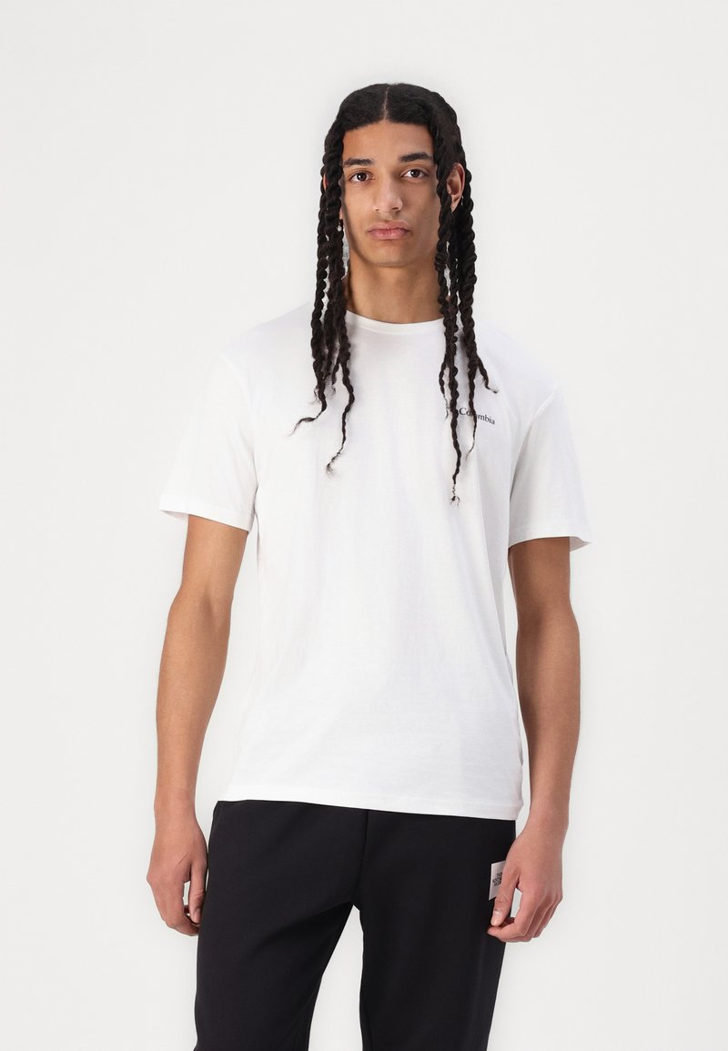 T-shirt en coton blanc à manches courtes, col rond et détail de logo sur la poitrine. Porté avec un pantalon noir.