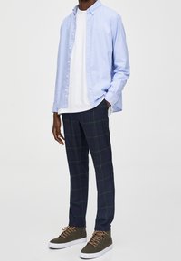 Hellblaues Hemd über einem weißen T-Shirt, kombiniert mit marineblauen karierten Hosen mit grünen Linien. Braune High-Top-Sneaker mit weißen Sohlen.