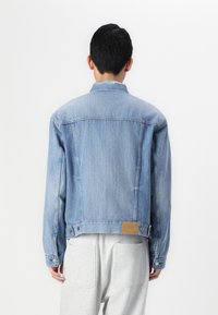 Lichtblauwe denimjas met een klassiek ontwerp, voorzien van een gespreide kraag, knoopmanchetten en een leren patch op de taille aan de achterkant.