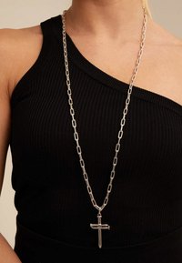 Collar de cadena de plata con eslabones alargados y un colgante de cruz, llevado sobre una blusa negra con ribetes que presenta un escote asimétrico.