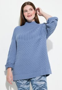 Ljusblå stickad tröja med hög krage, strukturerade mönster och ribbade muddar. Buren över mönstrade blå leggings.