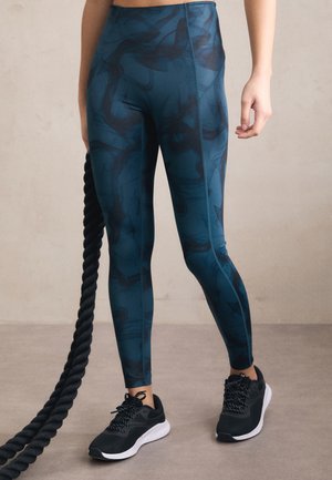Leggings made of stretchy blue fabric with a dark abstract pattern, paired with black athletic shoes featuring white soles and textured laces.