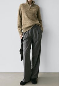 Pull marron en maille torsadée avec col montant, associé à un pantalon gris plissé et des mocassins noirs. Sac à main foncé tenu à la main.