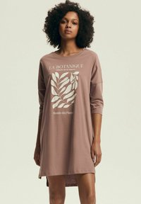 Kobieta o kręconych włosach, ubrana w luźną sukienkę w kolorze mauve z białym roślinnym wzorem i francuskim tekstem na przodzie, stojąca na jednolitym tle.