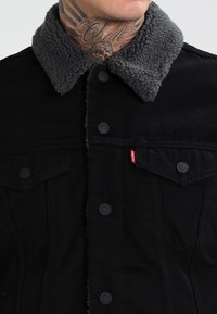 Tatuaggio sul collo di un uomo visibile sopra una giacca nera con bottoni e colletto in pile grigio, con etichetta rossa sulla tasca del petto.