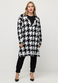 Zwart-wit houndstooth-patroon lange cardigan met een revers, knoopsluiting en zijsplitten, gecombineerd met zwarte broek en hakken.