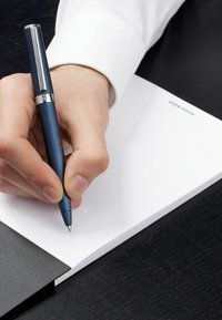 Sininen metallikynä pidetään valkoisen muistikirjan päällä, jonka kansi on mustaa texturea; käsi, jossa on hienoja karvoja, on valmiina kirjoittamaan.