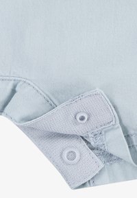 Ljusblå bomullsplagg med volangdetalj och tryckknappar. Har släta sömmar och mjuk textur, lämplig för bekväm användning.