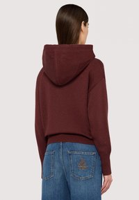 Burgundy hoodie med ribbat stickad textur och bred krage, kombinerad med blå jeans med broderad design på bakfickan.