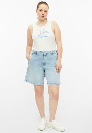 Femme portant un haut blanc sans manches avec un motif plage et un short en jean bleu clair, debout les mains dans les poches.