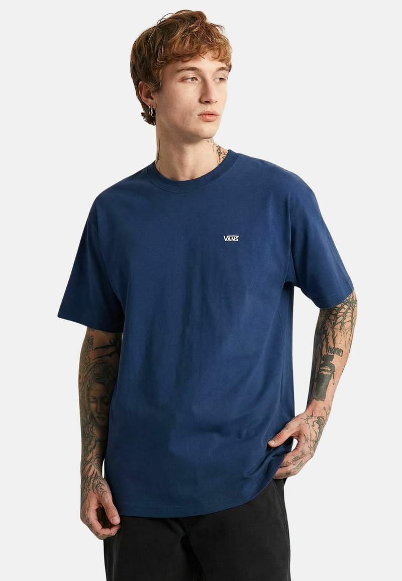 Jeune homme aux cheveux roux et tatouages portant un T-shirt Vans bleu marine, regardant sur le côté avec une main dans la poche et l'autre tenant l'ourlet du T-shirt.