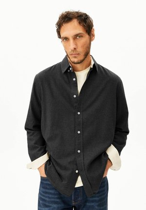 Mann mit kurzen lockigen Haaren, trägt ein dunkles, geknöpftes Hemd über einem hellen Langarmhemd und blaue Jeans, Hände in den Taschen, neutraler Gesichtsausdruck.