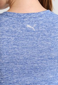 Acercamiento a una camiseta deportiva de tela azul jaspeada en la espalda de una persona, con un pequeño logo de un gato saltando en blanco cerca del escote.