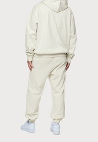 Persona di spalle, indossando un felpa beige con cappuccio e pantaloni della tuta abbinati, completati da sneakers bianche, su uno sfondo bianco semplice.