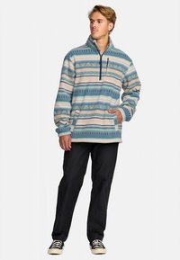 Pulover od flisa u svijetloplavoj, krem i zelenoj boji s geometrijskim uzorcima, ovratnikom s pola zatvarača, bočnim džepovima i opuštenim krojem, uparen s crnim hlačama.