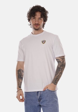 Uomo con capelli ricci e tatuaggi su entrambi gli avambracci, che indossa una maglietta bianca con un piccolo logo e jeans blu, con una mano in tasca.