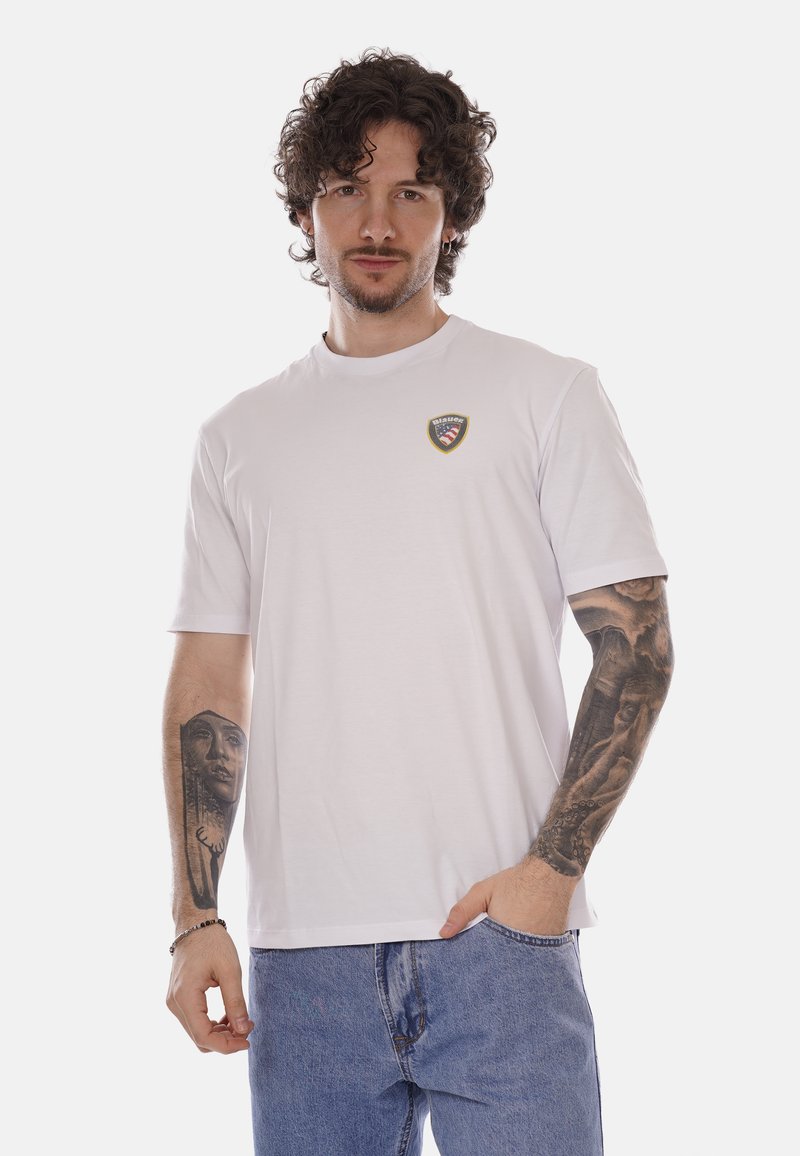 Uomo con capelli ricci e tatuaggi su entrambi gli avambracci, che indossa una maglietta bianca con un piccolo logo e jeans blu, con una mano in tasca.