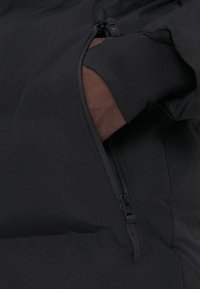 Χέρι με μαύρο γάντι μέσα σε μερικώς ανοιχτή τσέπη μαύρου ρούχου που δείχνει λεπτομέρειες φερμουάρ και υφή υφάσματος.