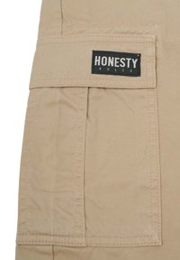 Beige bomuldsstof med en lappelomme. Mærket står "HONESTY" med hvid tekst på en sort baggrund. Enkel syning og holdbare design detaljer.