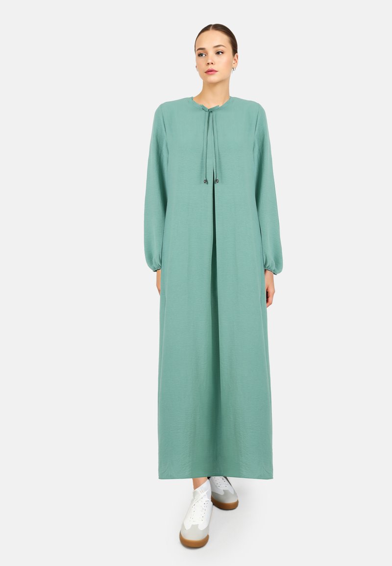 Long, mint green dress with a round neck, drawstring closure, and puffed sleeves. Worn with white sneakers featuring brown soles.