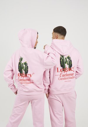 Różowe bluzy z kapturem i spodnie, z grafiką kaktusa z tyłu, na której widnieją słowa "Legacy" oraz "Cactaceae Curation Studios."