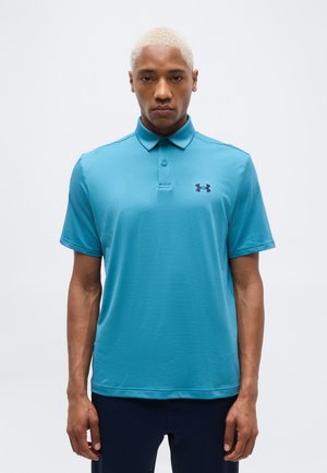 Homme aux cheveux blonds portant un polo bleu Under Armour et un pantalon foncé, debout devant un fond blanc uni.