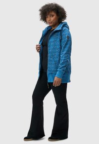 Veste à capuche zippée bleu à motifs, avec une capuche, des poches frontales et des poignets côtelés, portée sur un t-shirt noir et assortie avec un pantalon noir évasé.