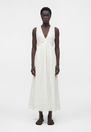 Kobieta o krótkich kręconych włosach, ubrana w białą, długą suknię bez rękawów z dekoltem w serek i brązowe sandały, stoi na jednolitym tle.