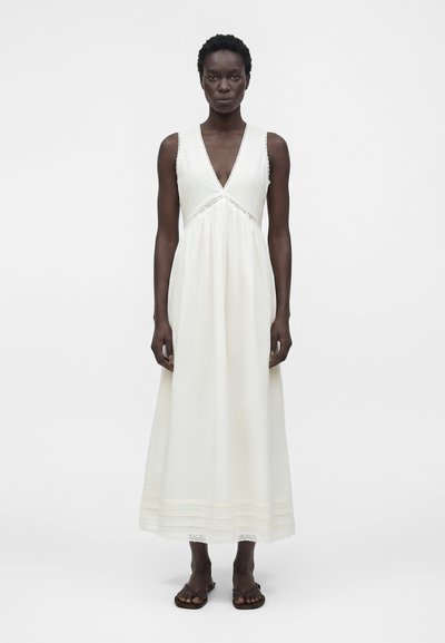 Woman with short curly hair wearing a sleeveless white maxi dress with a V-neck and brown sandals, standing against a plain background.