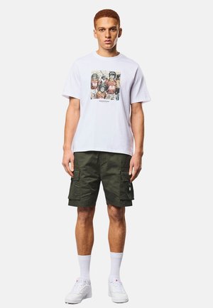 Junger Mann mit kurzen roten Haaren, der ein weißes T-Shirt mit Grafikdruck, dunkelgrüne Cargoshorts, weiße Socken und weiße Turnschuhe trägt.