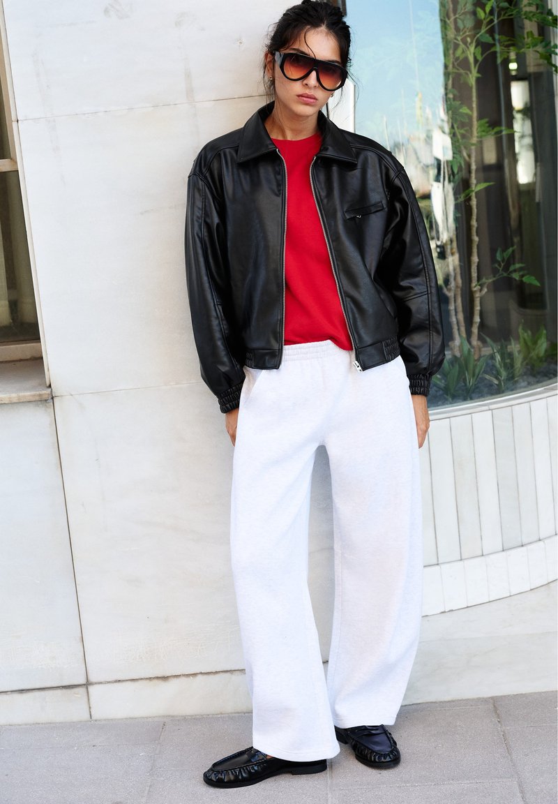 Femme portant une veste en cuir noire oversize, une chemise rouge, un pantalon blanc ample, des chaussures noires et de grandes lunettes de soleil, debout contre un mur clair.