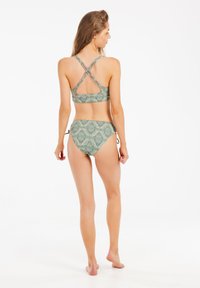 Komplet bikini z zielonym wzorem geometrycznym. Góra ma krzyżujące się ramiączka i detal w kształcie dziurki; dół jest wiązany po bokach. Gładka struktura.